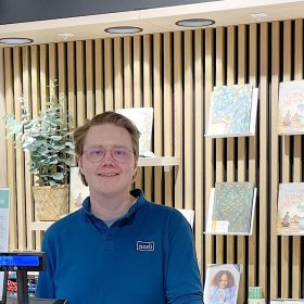 En mann i med briller og blå skjorte står foran en vegg med noen bøker.