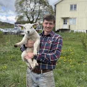 En mann i rutete skjorte og arbeidsbukse står ute på et jorde og holder et lam i armene. Han smiler mot kamera. I bakgrunnen er det gress, trær, biler og et lyst gårdshus under en skyet himmel.