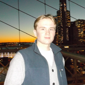 En mann med blondt hår, hvit skjorte og grå vest står foran høye bygninger i en stor by