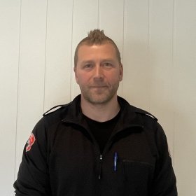 En mann med svart uniformsjakke og lyst hår og skjegg står foran en lys vegg.