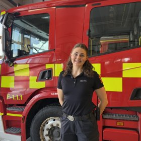 En kvinne i mørk uniform står foran en stor, rød brannbil inne i en brannstasjon. Hun har mørkt, krøllete hår trukket bakover og smiler mot kamera. På seg har hun svart poloskjorte, mørk bukse, belte med spenne og samband på hoften. Brannbilen bak henne har gule refleksfelt og store hjul som fyller deler av bakgrunnen.