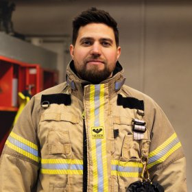 En mann med mørkt hår og skjegg. Han har på seg utrykningsdrakt. Han er inne.