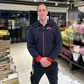 En mann med kort hår og mørk hettegenser står foran et blomstertorg med rosa og gule blomster. 
