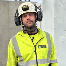 En byggarbeider med litt skjegg står utendørs på en byggeplass. Han har på seg en gul refleksjakke med logoen «Con Form» på brystet, og en hvit hjelm med hodelykt og hørselsvern. Han ser rett mot kameraet og smiler svakt. Bak ham sees en grå betongvegg og deler av en hvit innpakning.