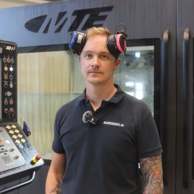 En person står foran en stor CNC-maskin med kontrollpanel. Han ser inn i kameraet og har hørselsvern på hodet. CNC-maskinen bak dem har store vinduer og kontrollskjerm med knapper og måleindikatorer. Personen har på mørkeblå arbeidsklær og tatoveringer på armen.