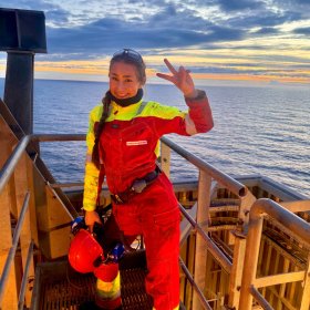 En ung kvinne i rødt offshorearbeidstøy står på en plattform med havet og en dramatisk solnedgang i bakgrunnen. Hun smiler mot kameraet og holder opp et fredstegn med hånden, mens hun holder vernehjelmen i den andre.