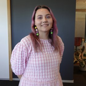 Jente med rosa hår og rosa og hvit rutete kjole som smiler foran en vegg.