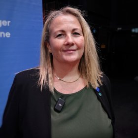 Kvinne med lyst hår i svart dressjakke, grønn bluse og gullsmykke smiler rett frem til kamera med blå bakgrunn.