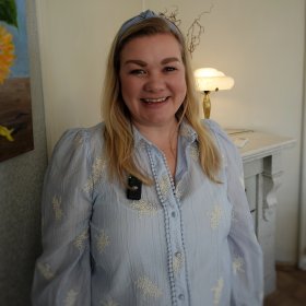 Kvinne med blondt hår og blå bluse med hårbølje. Hun smiler til kamera med et bilde på veggen ved siden av.