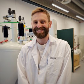 Mann med hvit frakk og brunt hår smiler til kamera. Han står foran et hvitt sylinderformet apparat 