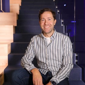 DevOps-utvikler Eirik Luka på jobb.  En mann sitter avslappet på en mørk trapp innendørs, med et varmt smil mot kameraet. Han har på seg en lys, stripete skjorte og mørke olabukser. Bak ham skimtes moderne kontorinteriør med blått lys og glassrekkverk.