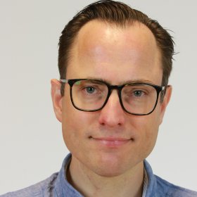 Forsker En mann med briller og mørkt hår, gredd bakover, ser rett i kamera. Han har på seg en lyseblå skjorte med ståkrage og hvit bakgrunn. Bildet er tatt fra brystet og opp.