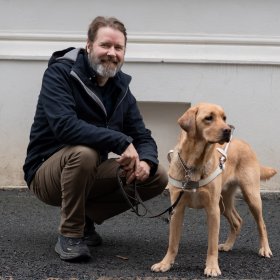 En mann i mørke klær. Han smiler og ser rett i kamera. Han sitter på huk og holder en førerhund i bånd. Det er en lys labrador. De er ute.