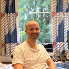 En mann sitter på et kontor og smiler mot kameraet. Han har på seg en lys T-skjorte og sitter ved et skrivebord med PC og papirer. Bak ham er et stort vindu med utsikt mot grønne trær og mønstrede gardiner på hver side.