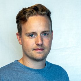 En mann med blondt hår og blå genser står foran en lys vegg.