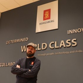 En mann med mørkt langt skjedd, caps og blå langermet genser står med armene i kors foran brystet og ser inn i kamera. Han står foran en vegg. 