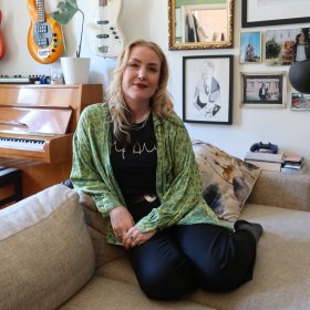 En dame i grønn skjorte og mørke bukser. Hun sitter smilende i en sofa i en personlig og fargerik stue. Bak henne henger flere gitarer på veggen, og et piano står ved siden av en stol. Veggen er dekorert med bilder og kunst i ulike rammer.