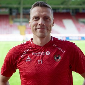 En mann i rød fotballdrakt. Han smiler og ser rett i kameraet. I bakgrunnen er en fotballbane og en tribune.