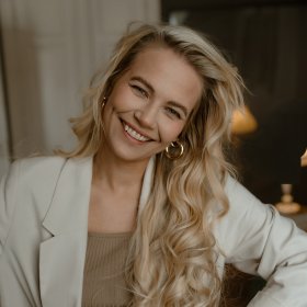 Kvinne med langt lys hår smiler til kamera. Hun har på seg hvit blazer og beige topp. 
