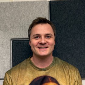 En mann med kort mørkt hår står vendt rett mot kameraet og smiler svakt. Bak ham er grå og mørke akustiske lydpaneler montert på veggen.