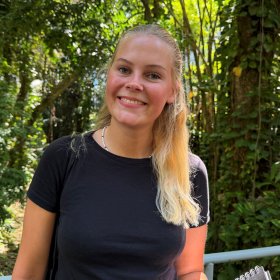 Kvinne med langt blondt hår iført svart T-skjorte og blått jeansskjørt poserer og smiler mot kamera. I bakgrunnen ser man trær og grønt.