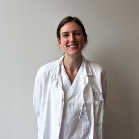 En kvinne med mørkt hår står i hvit frakk foran en hvit vegg