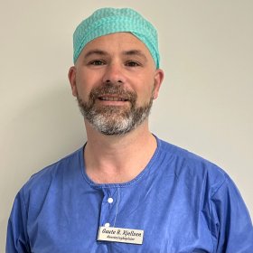 En mann med skjegg står inntil en hvit vegg. Han har på seg blå medisinsk overdel og blå hette på hodet. Han smiler og han har på et navneskilt på brystet på overdelen/skjorten.