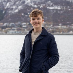 En mann i lys genser og mørkeblå jakke. Han ser rett i kameraet og smiler. Han er ute, bak han er havet, hus og fjell.