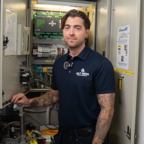 En mann står foran et elektrisk skap og smiler til kamera. Han har brunt hår og mange tatoveringer på armene. Han har på en mørkeblå t skjorte der det står "ALT heis"