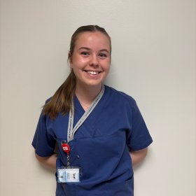 En ung kvinne med brunt hår i hestehale og blå skjorte står foran en lys vegg.