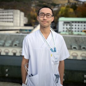 En mann i hvit legeuniform med mørkt kort hår, som står utendørs foran sykehusbygninger med fjell i bakgrunnen. Han ser rolig og profesjonell ut, med stetoskop i lommen og ID-bånd rundt halsen.