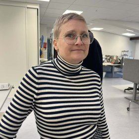 Korthåret dame med briller og stripete genser ser inn i kamera. Hun står i et hvitt rom med pulter og symaskiner i bakgrunnen.