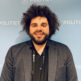 Mann med langt, mørkt og krøllete hår og langt skjegg står og smiler i et portrettbilde. Han har på seg svart skjorte og mørkegrå dressjakke. I bakgrunnen sees en lyseblå vegg med politilogo.