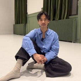 Mann med svart hår, blå skjorte, svarte jeans og ullsokker sitter på gulvet og ser mot kamera. I bakgrunnen er det et stort, åpent rom med hvitt gulv.