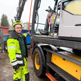 Anleggsmaskinfører i neon arbeidsklær og hjelm står ved en gravemaskin og poserer.