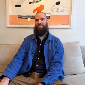 Mann med kort hår og langt mørkt skjegg, iført lyseblå jakke og mørkeblå skjorte, sitter i en sofa og poserer. I bakgrunnen henger en illustrasjon på veggen.