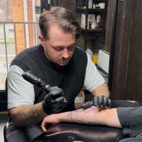 Mann med mørkt hår, hvit T-skjorte og svart vest holder en tatovørmaskin og gjør seg klar til å tatovere en kunde som ligger med armen frem. I bakgrunnen sees tatovørstudioet.