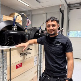 Mann med kort svart hår, svart arbeids T-skjorte og vernebukse står ved ferdigstilte produkter som er klargjort for utsending til kunder.
