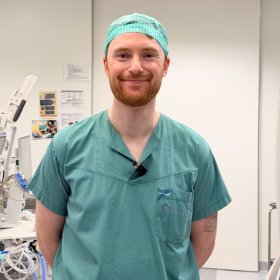 Mann med oransje skjegg iført grønt kirurgisk arbeidstøy og kirurgisk hette står og smiler mot kamera.