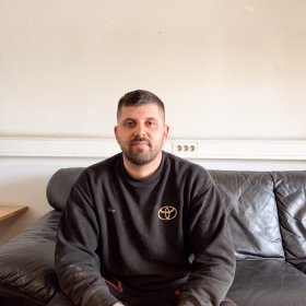 Mann med kort svart hår, svart genser og svart arbeidsbukse sitter i en skinnsofa i et pauserom på arbeidsplassen og poserer til et bilde.