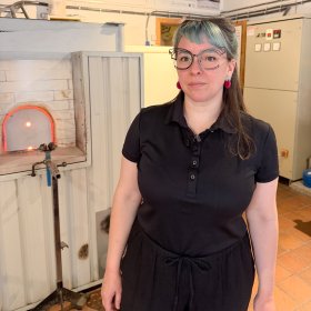 Kvinne med langt mørkt hår og turkis pannelugg, iført briller med klart glass, svart T-skjorte og svart bukse, står og poserer til portrett i et glassblåserverksted. I bakgrunnen sees utstyr og arbeidsmiljø fra verkstedet.