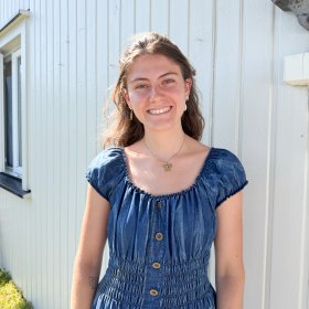 Kvinne med langt brunt krøllete hår står foran kamera og smiler. Hun har på seg en blå denimkjole, og i bakgrunnen vises en lys vegg og grønt gress.