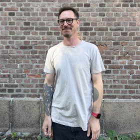 Mann med lys grå t-skjorte og briller står foran en murvegg og smiler.