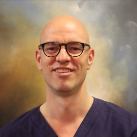 En mann smiler til kameraet. Bakgrunnen er et bilde klinikken har på veggen.  