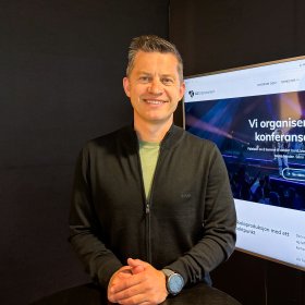 Mann med kort, mørkt hår smiler mot kamera. Han har på seg svart genser med en lys t-skjorte synlig i åpningen. En TV står på i bakgrunnen, inne på kontoret hans.