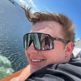 En mann med kort hår og solbriller og svart trøye med blå himmel bak.ak