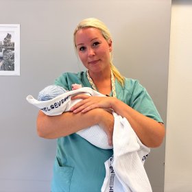 Barnepleier Andrea Selnes Otten Kvinne med langt blondt hår samlet i strikk bak, iført turkis/grønn arbeidsuniform, holder en nyfødt baby pakket i hvit pledd foran kamera.