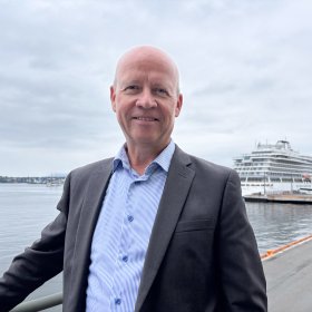 Mann i mørk grå dressjakke og blå skjorte står foran kamera. I bakgrunnen ses et cruiseskip og havet under overskyet himmel.