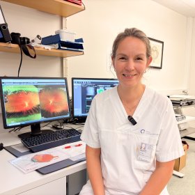 Portrettbilde av Øyelege Sigrid de Rodez Benavent Kvinne med lyst hår i strikk, iført hvit arbeidstøy, sitter på et kontor. Hun smiler og ser inn i kamera. Bak henne står to PC-skjermer og trykksaker fra et øyelegekontor.