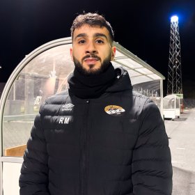 Mann med svart hår, skjegg og bart poserer foran en fotballbås utendørs om kvelden. Han har på seg en langermet jakke, og lys fra stolper skimtes i bakgrunnen.